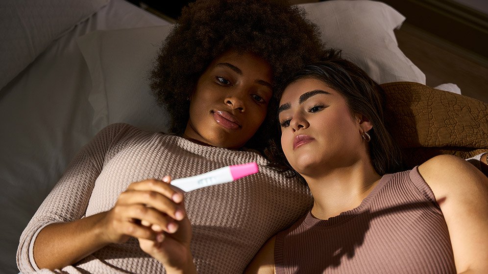 Two women lying in bed, both holding a pregnancy test and looking at it.