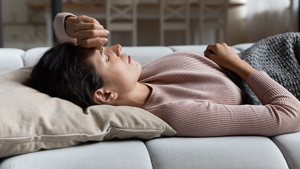 A woman lying on the couch with a blanket, her hand over her head, looking tired or fatigued.