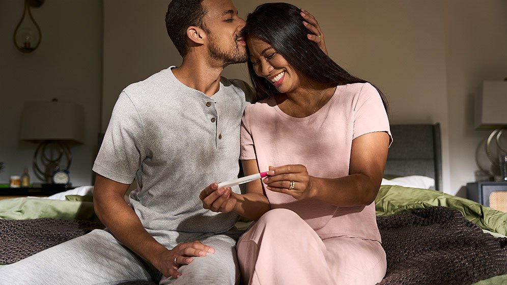 A woman and man sitting on the edge of the bed, with the woman excitedly holding a pregnancy test and the man kissing her forehead.